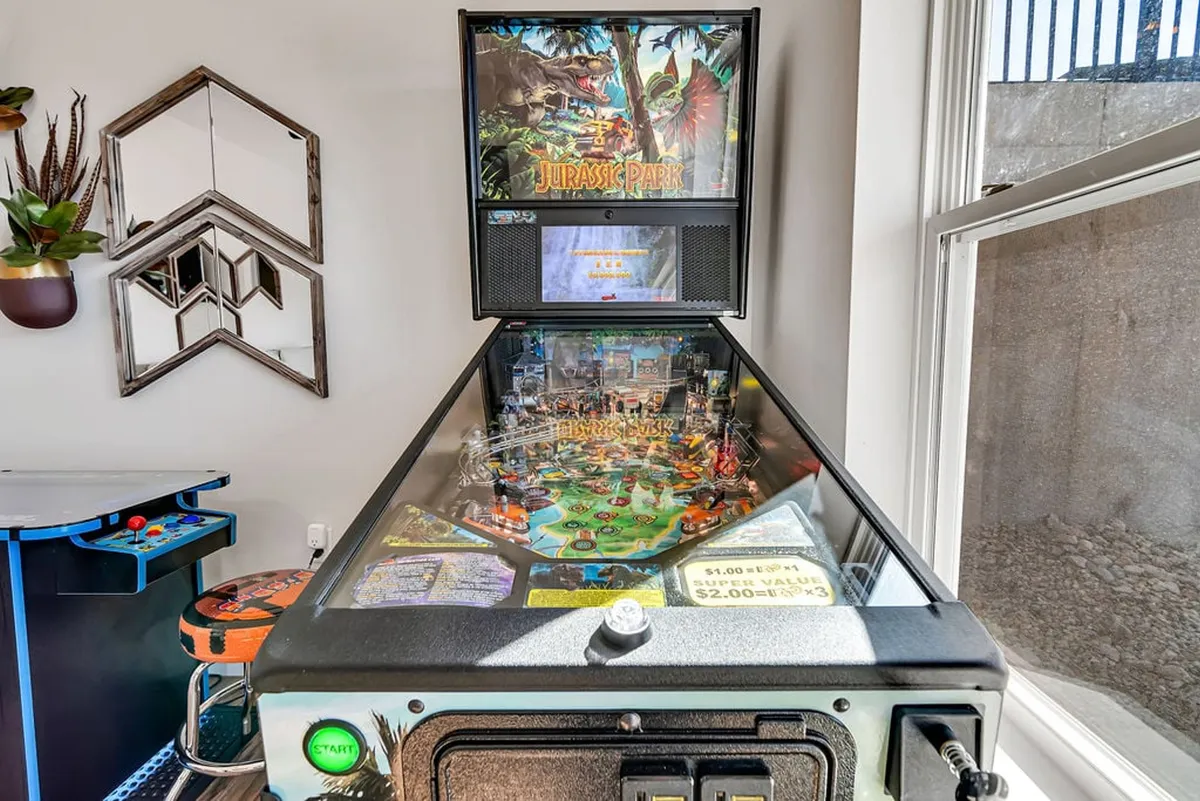An interior view of a game room featuring a Jurassic Park pinball machine, geometric wall mirror, and another arcade game. The space exudes a fun, nostalgic vibe, inviting both kids and teens for friendly competitions and entertainment when outdoor play isn't possible.