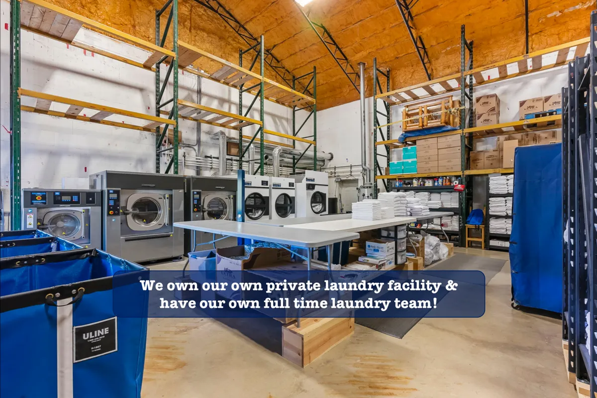 An interior scene showcasing industrial laundry machines, organized laundry baskets, and storage shelves. The efficient, spotless setup is clearly designed for professional laundering of linens and towels.