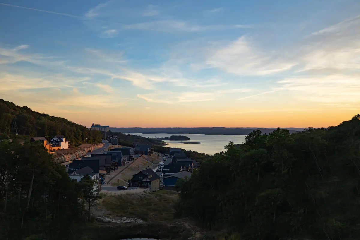 A panoramic view of the lake at sunset, with houses and trees along the water's edge. The scene emanates calm, with a glowing sky and the promise of lakeside relaxation, boating, or peaceful evening strolls.