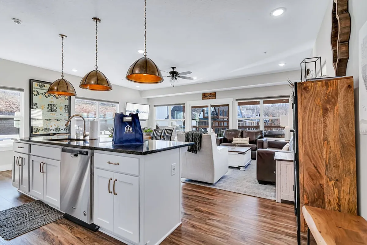 This image captures a spacious open-plan area that combines a kitchen island with bar stools, stylish hanging pendant lights, a cozy living room seating area, and large windows that flood the space with natural light. The kitchen is fully equipped and seamlessly flows into the living and dining areas, showing off communal spaces where guests can cook, dine, and relax together.