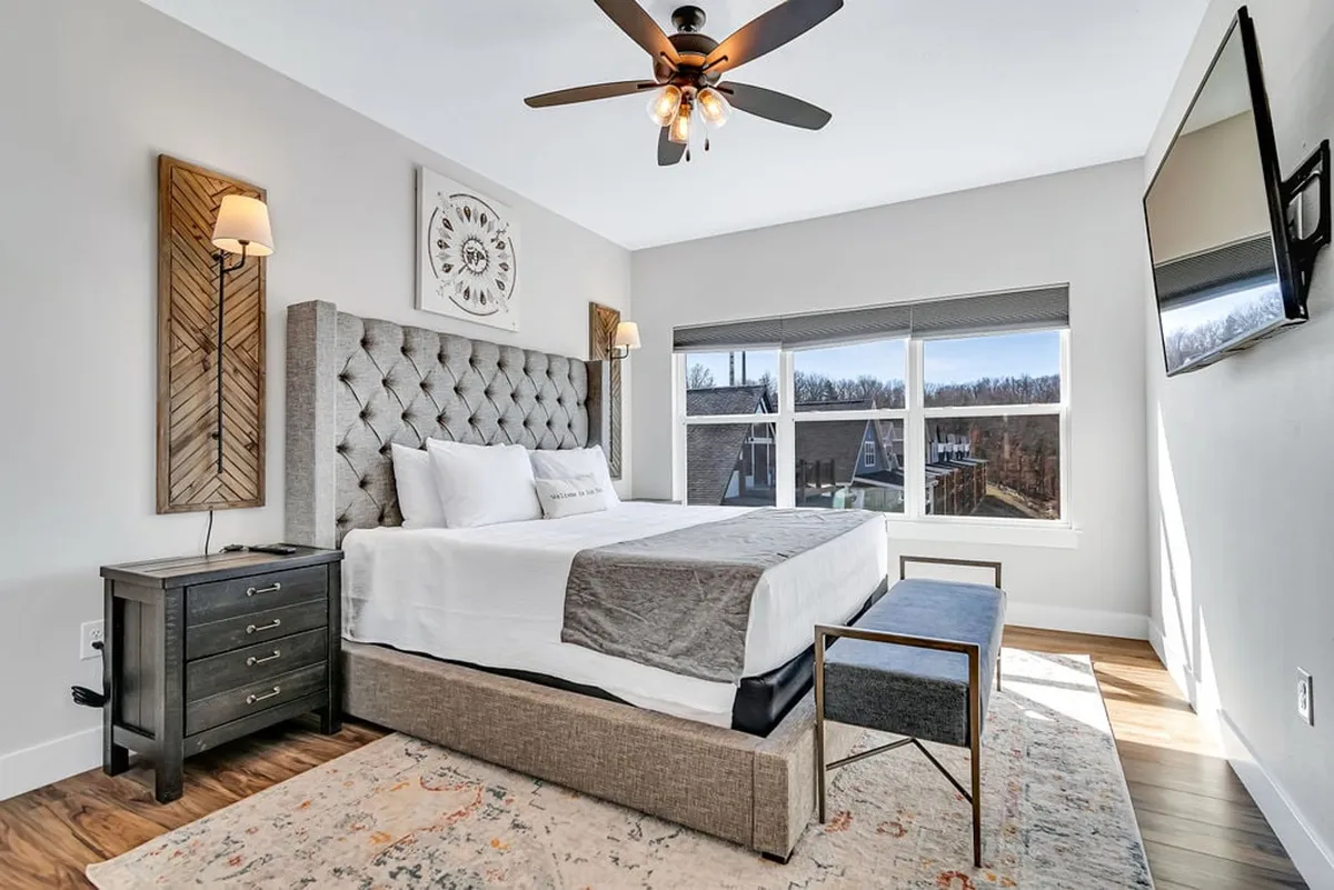 A beautifully appointed King bedroom featuring a tufted headboard, crisp white linens, large windows overlooking a scenic view, tasteful decorative wall art, and a nightstand with a reading lamp. The room exudes a sense of light-filled serenity and comfort, inviting guests to relax and unwind.