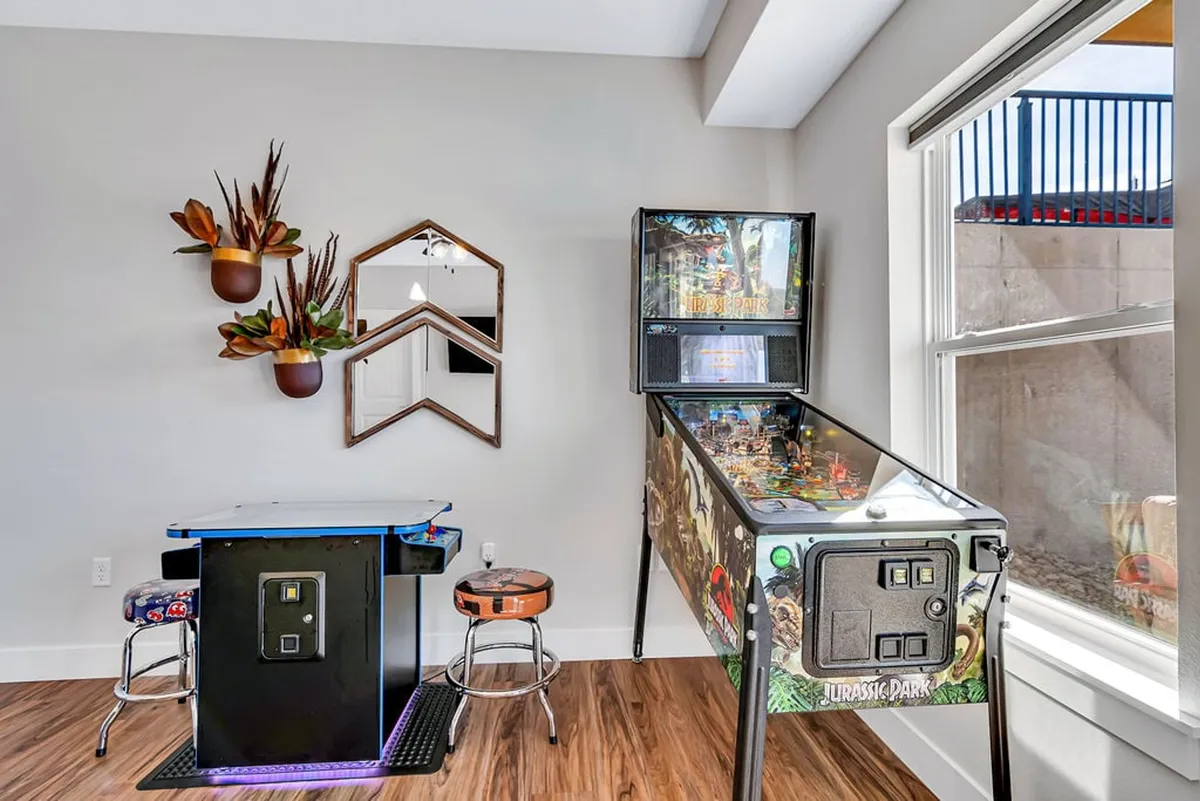This image features a lively game room with a Guardians of the Galaxy Pro Pinball Machine, a game table, decorative plants, and a mirror. The space is well-lit and inviting, geared towards socializing and playful competition.