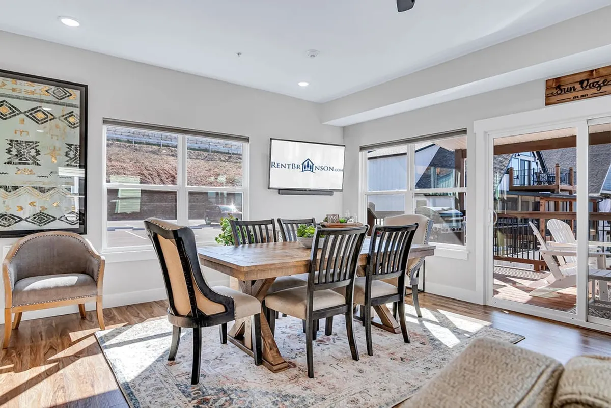 This image features a spacious dining table surrounded by comfortable chairs, set near large windows and accented with decorative wall art. A TV screen is visible in the background, emphasizing the dining area's connection to the living space. The warm, inviting atmosphere suggests opportunities for group meals, game nights, or sharing stories.
