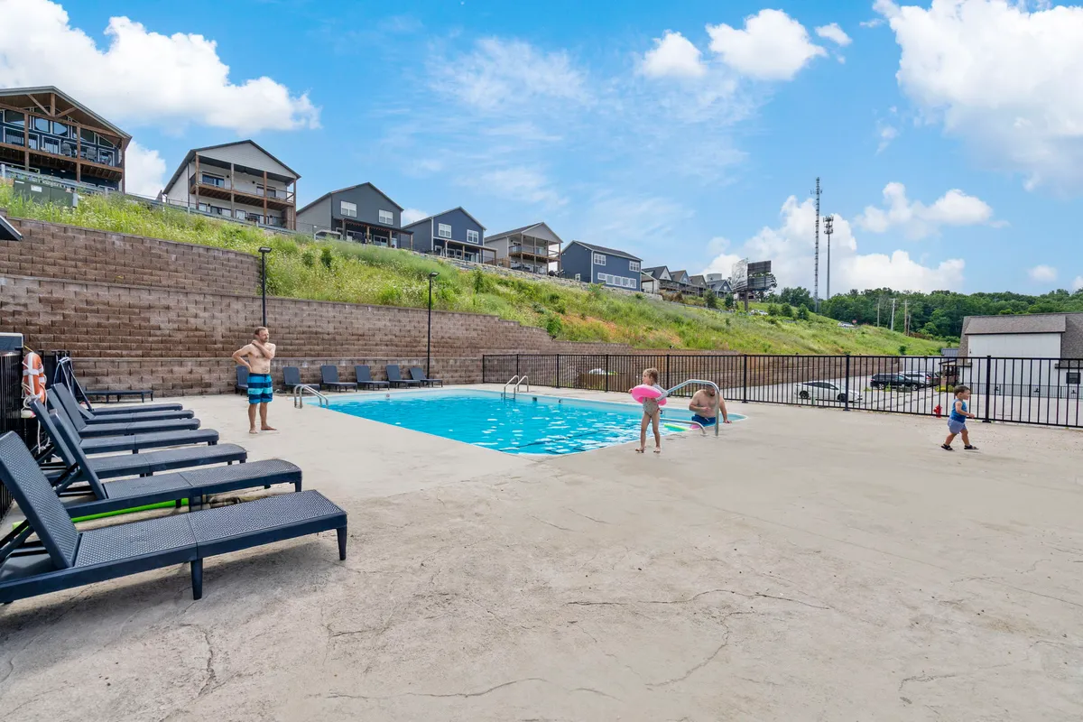 A large outdoor swimming pool is bustling with activity as children splash in the water and adults lounge nearby. Lounge chairs and shaded areas provide relaxation while families enjoy quality time together under the sun.