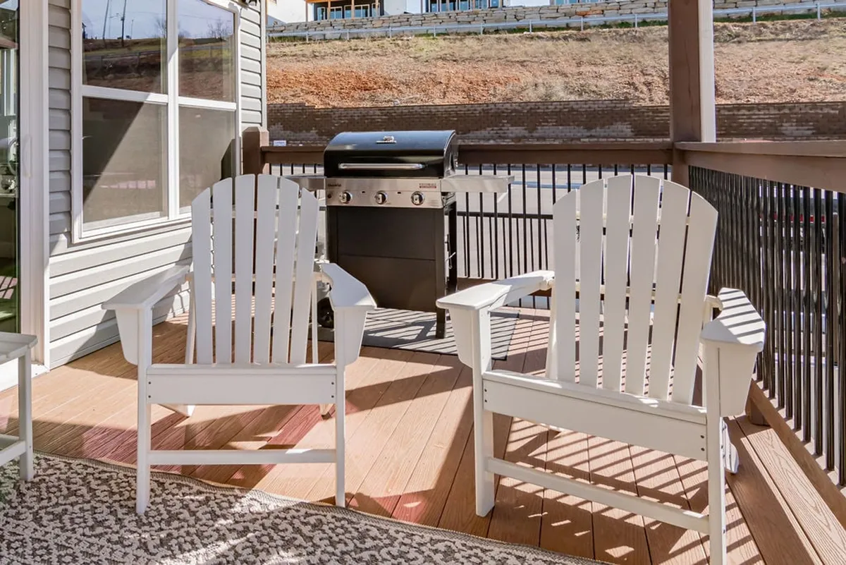 This image captures a spacious deck area featuring white Adirondack chairs, a grill, and ample room for gathering. The outdoor setting invites relaxation and socializing while overlooking natural surroundings, suggesting opportunity for sunset viewing and al fresco dining.