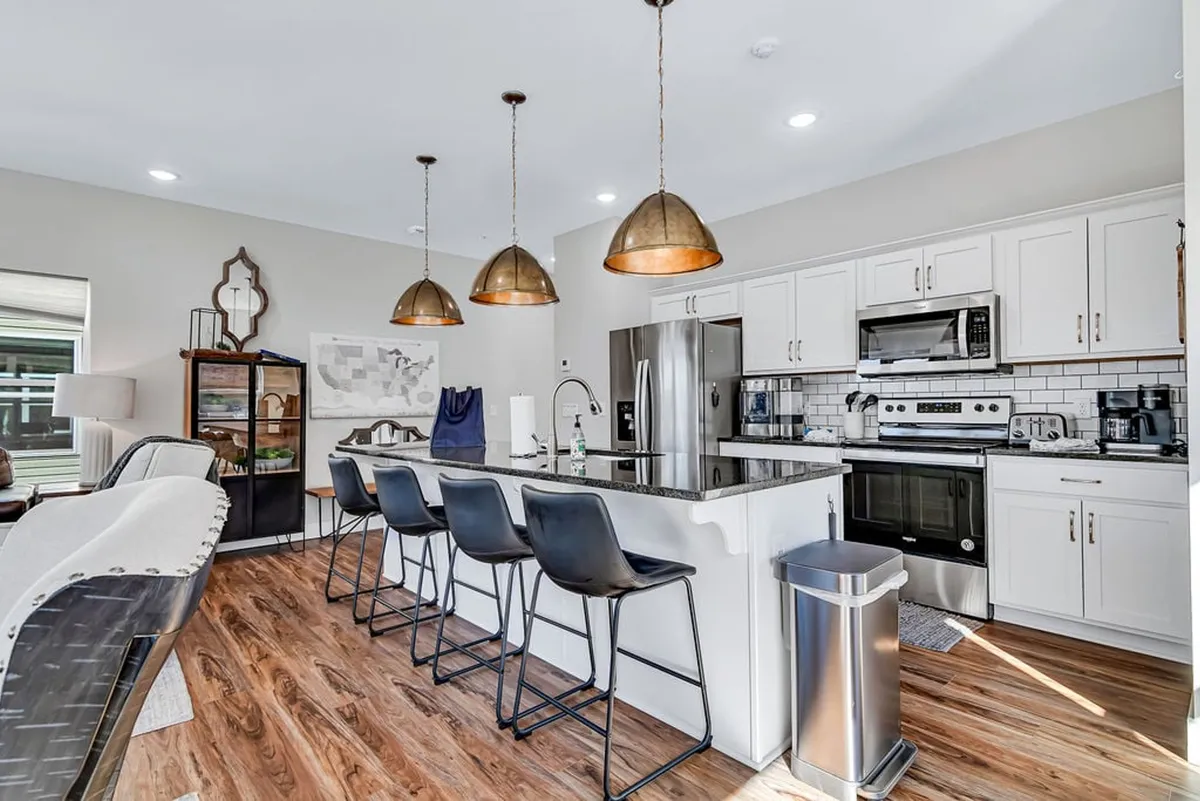 This image showcases a contemporary kitchen with a prominent island, bar stools, pendant lighting, and stainless steel appliances. The setup is ideal for cooking, socializing, and entertaining, and suggests a welcoming space for prepping meals or sharing snacks.