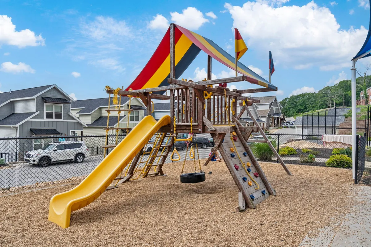 This image features a colorful playground structure with a canopy, slide, and swing set. Children are seen playing while parents supervise nearby, and the area appears lively and family-friendly. The playground is surrounded by green space, making it an inviting spot for family outings and playdates.